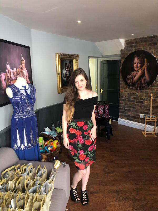 Clare Shapcott poses in red flowery skirt next to gold grand opening goody-bags with a blue dress and various wall displays around her.
