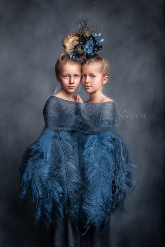 Two girls surrounded by blue material and large blue feathers wear a combined blue flower headpiece and touch hands.