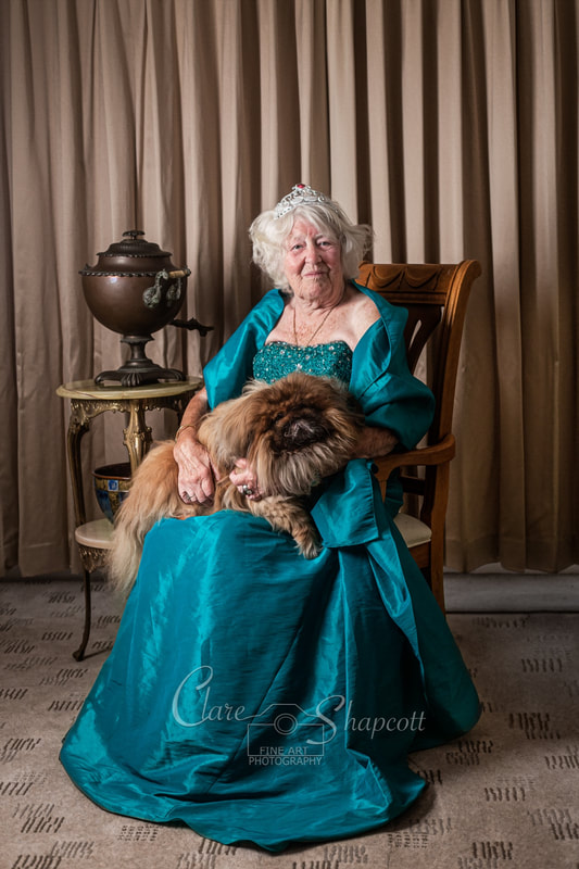 Elderly woman sits on chair in turquoise dress as she holds pekingese dog