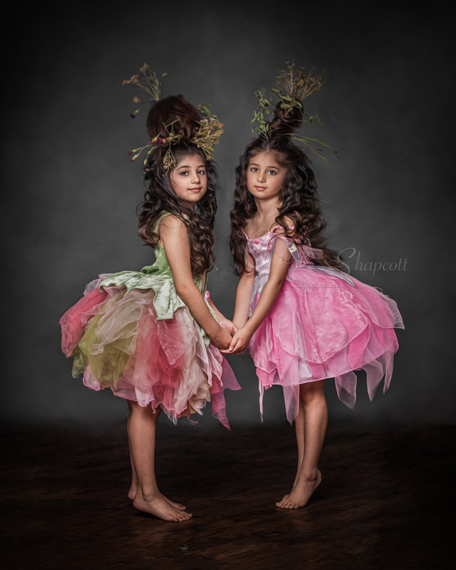 Two girls with tall, brown, wavy hair in pink and green tuille dresses hold hands and look at camera.