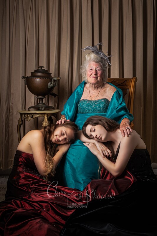 Grandmother sits royally on chair whilst two young women in long red dresses lay their heads asleep on her lap.