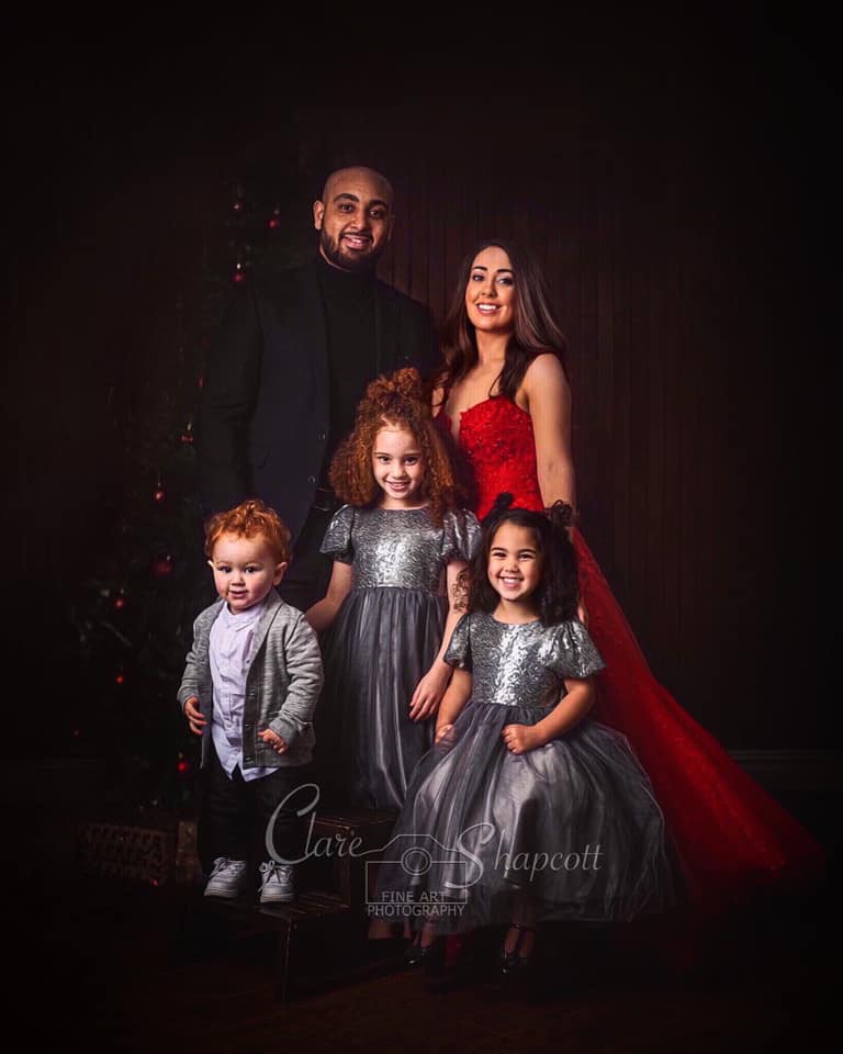 Lady in long red dress stands with her partner in a black suit and her 3 children in sparkly silver dresses and jacket.