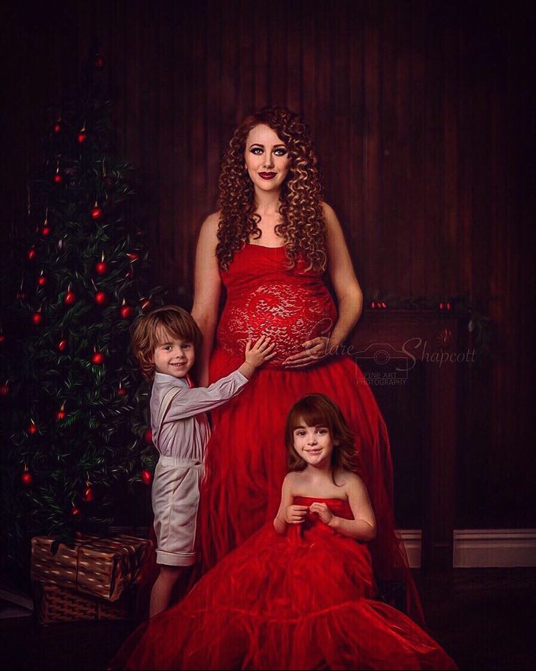 Pregnant lady with curly brown hair in red lace dress holds her bump as she stands with a boy in white shorts and a girl in a similar red dress with a christmas tree and presents next to them.