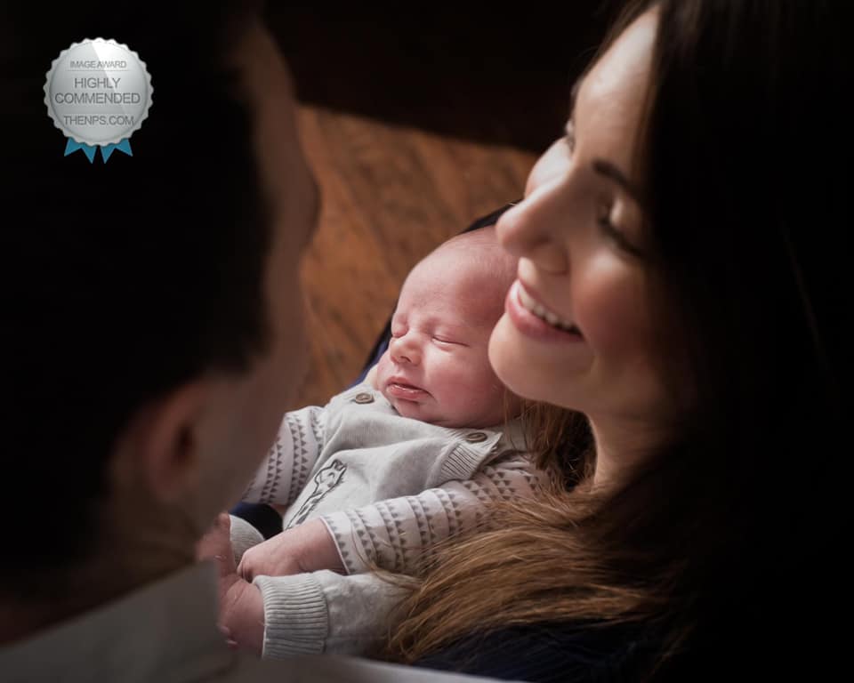 Mother and father smile at each other as photograph is taken over their shoulder of sleeping newborn baby.