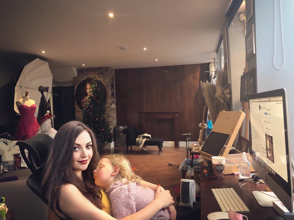 Behind the scenes photograph of Clare shapcott working at computer with young daughter asleep on her chest, Christmas tree in the background.