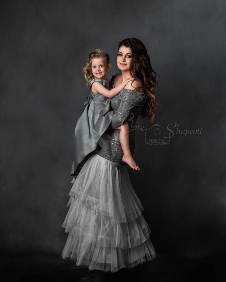 beautiful mum in silver tulle dress with wavy hair stands and holds daughter in grey dress.