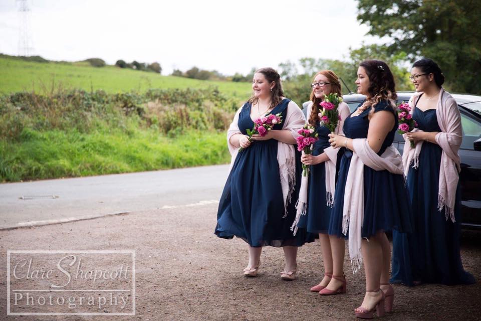 Bridesmaids await bride wearing matching navy blue dresses and pink shawls holding flowers