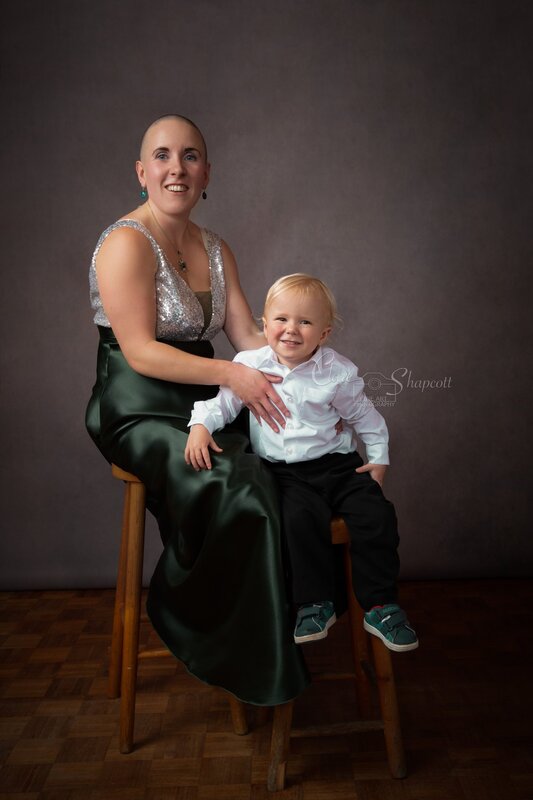 Woman in silver and green dress sits on wooden stool behind her smartly dress two year old son also sitting on a wooden stool.