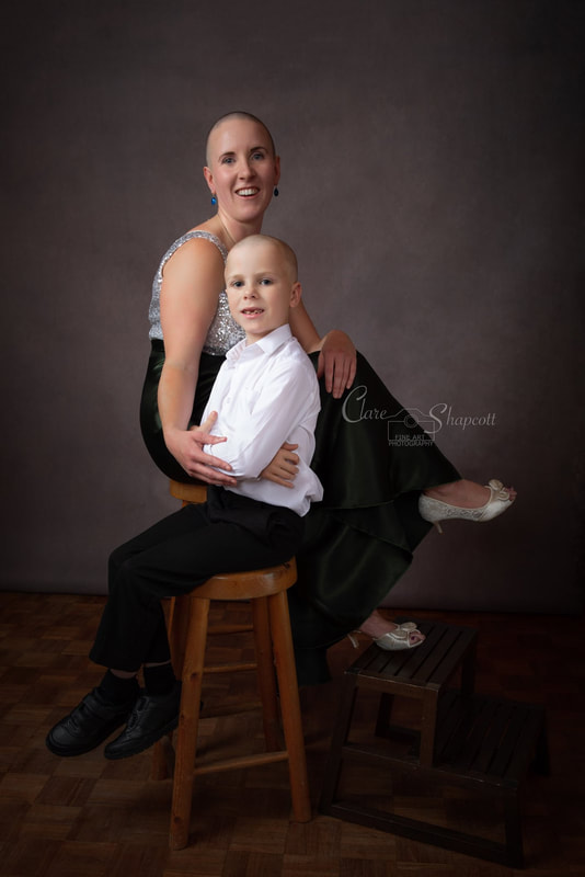 Woman in green and silver dress sits on a stool with her smartly dress son, both smiling and with shaved heads.