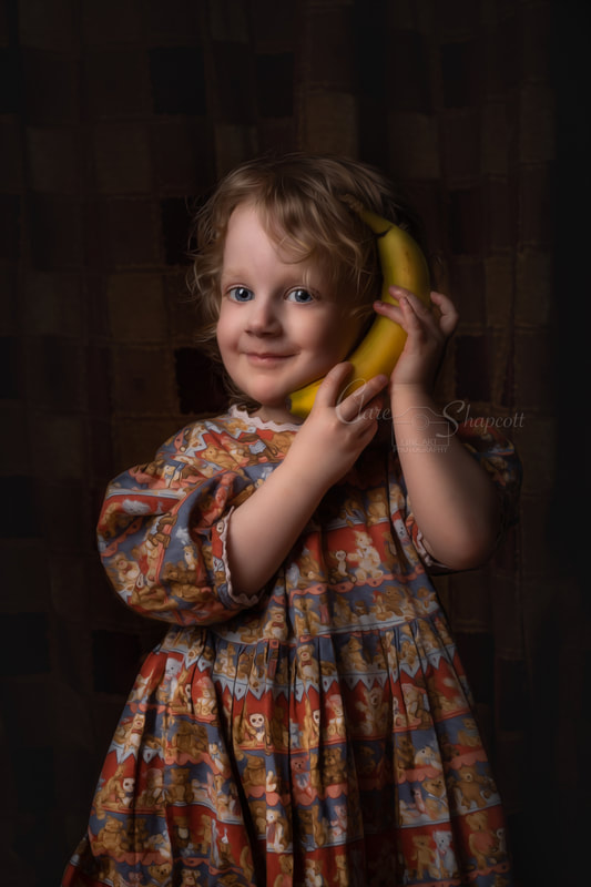 Fine art portrait of young girl in red and blue teddy bear dress holds banana to ear as a phone.