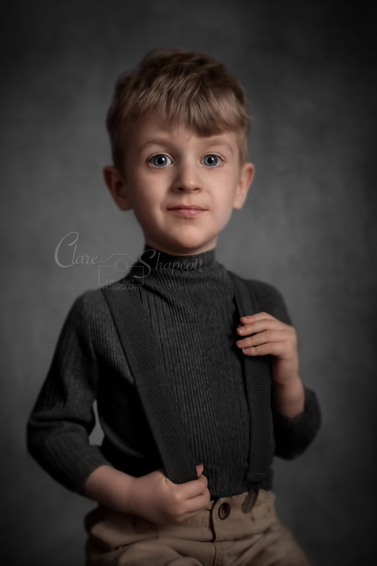 Young boy in grey turtleneck jumper holds suspenders in front of grey background.