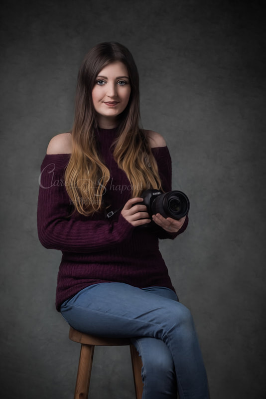 Woman in purple turtleneck jumper and blue jeans sits crossed legged on stool holding professional camera.