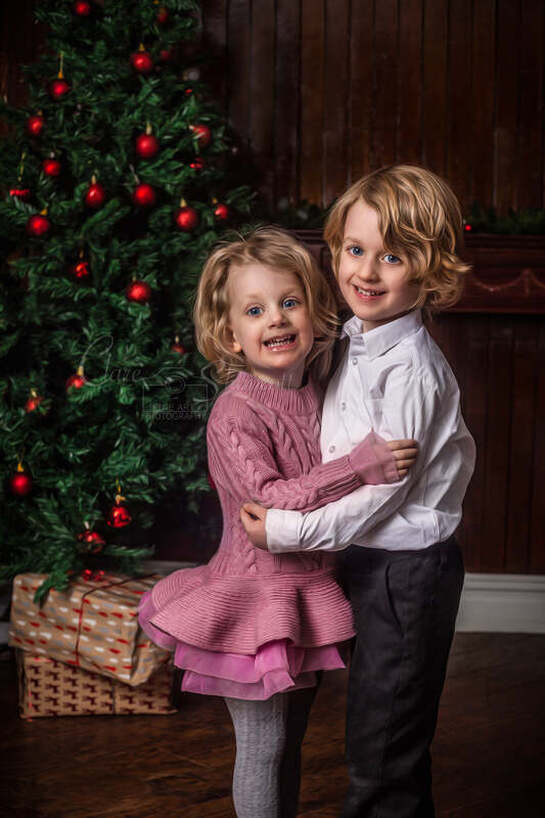 Portrait of brother and sister holding each other and smiling with a christmas tree and presents behind them.