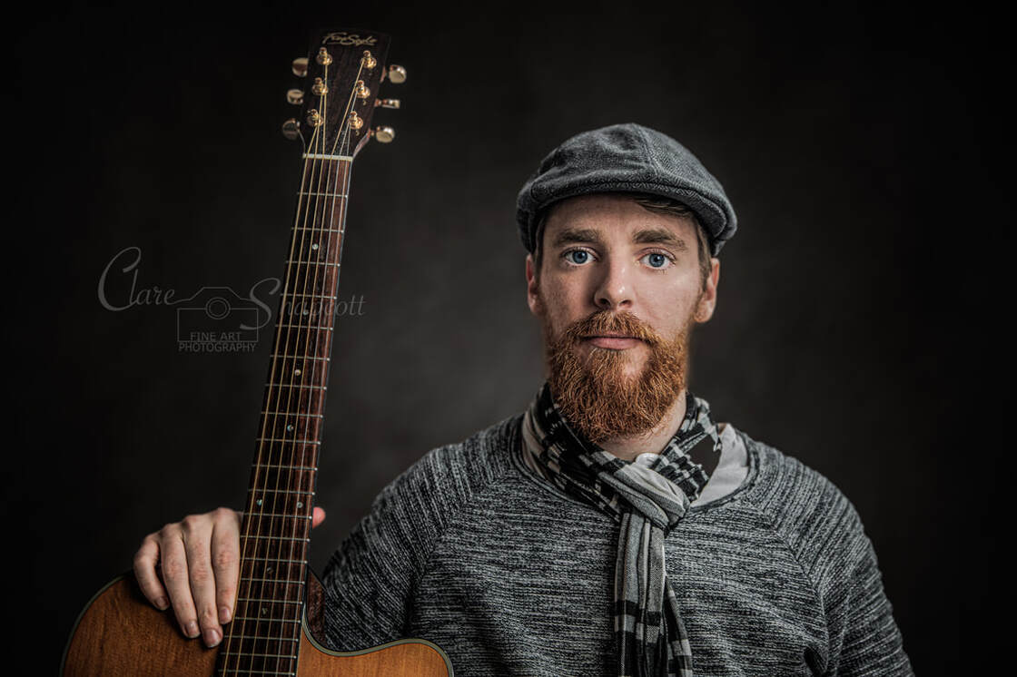 Fine art portrait of bearded musician with grey cap and scarf holding guitar upright.