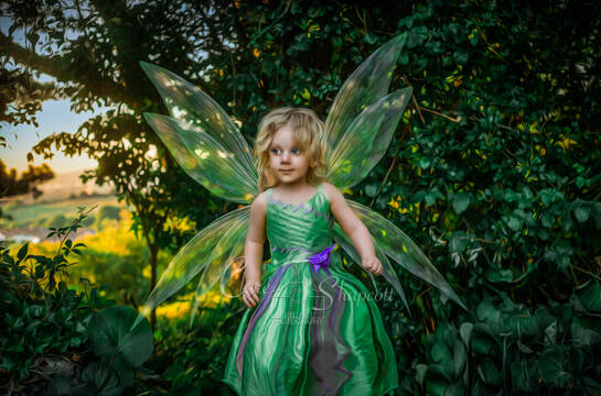 Young girl in purple and green dress with fairy wings stands in front of green bush and smiles.