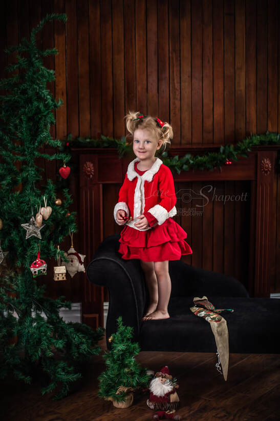 Little girl in red and white cardigan and red skirt standing on black chaise as she decorates a christmas tree