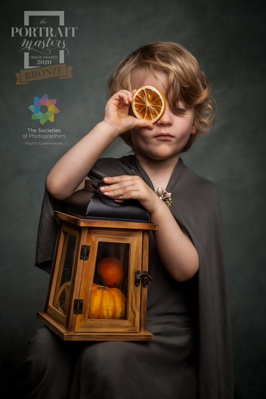 Fine art portrait of young boy in grey cloak holding lantern with small pumpkins inside, and also holding orange slice in front of eye.