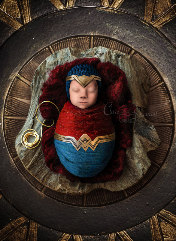 Wonder woman themed wrapped newborn lying down on top of red material with miniature lasso next to her.