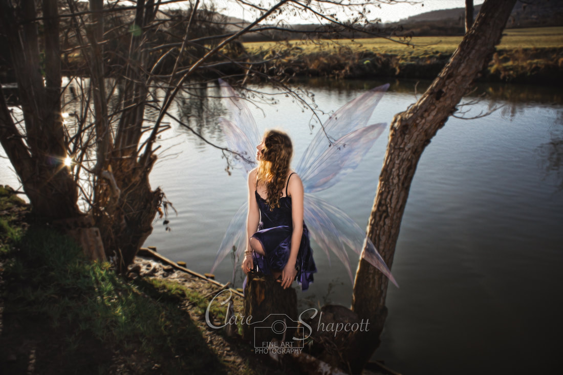 Illustrative photoshoot of young lady perched on top of tree stump with fairy wings in front of river.