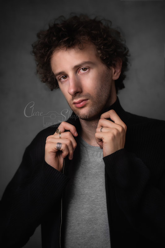 Handsome man with curly brown hair holds collar of black jacket and looks at camera.