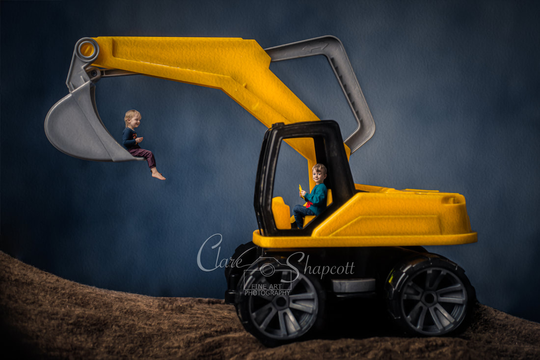 Young boy sits in his yellow toy digger holding steering wheel while young sister sits in digger bucket above the ground.