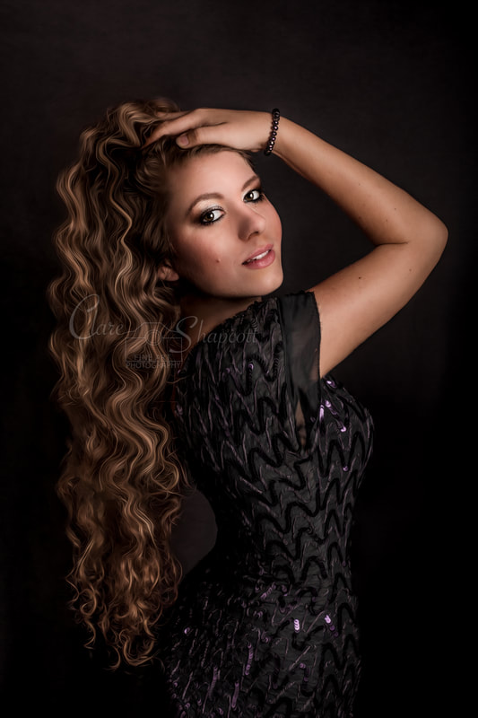 Beautiful lady in black dress and long wavy hair holds hands on head and looks at camera