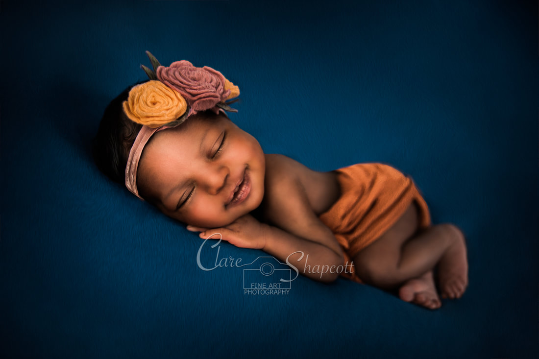 Newborn wrapped in orange material with a pink and orange flower headband sleeps face down on blue material.