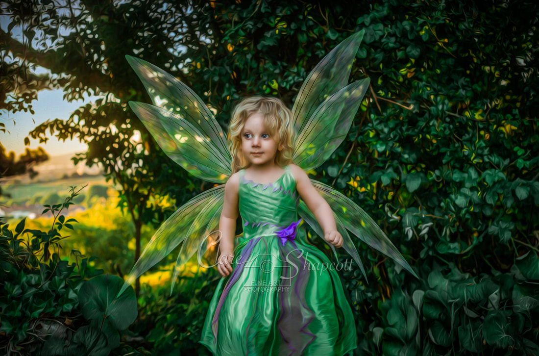 Young girl with purple and green dress with fairy wings stands in front of green bush and has a small smile.