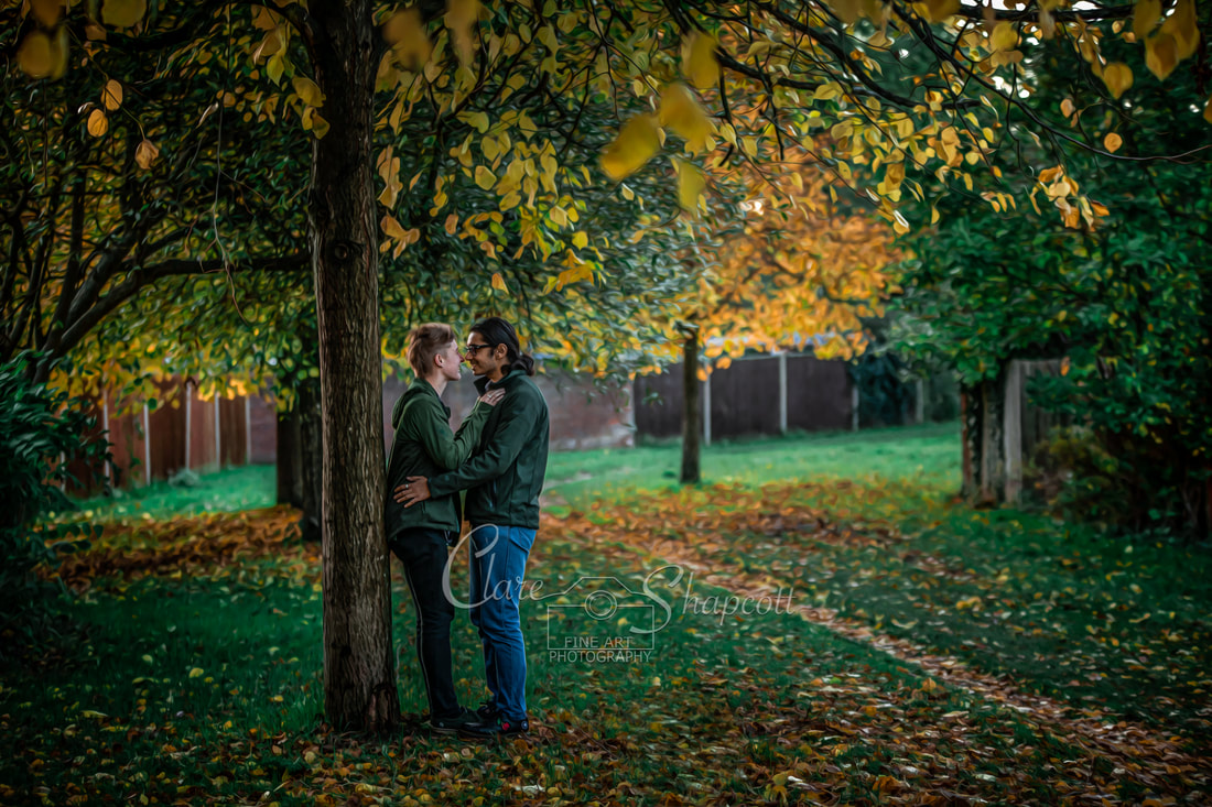 Young couple stand facing each other and leaning against a tree with yellow orange and green leaves all around them.