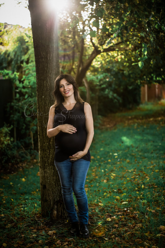 Pregnant woman in black top and blue jeans stands outside next to a tree with hands around bump and the sun shining through behind the tree