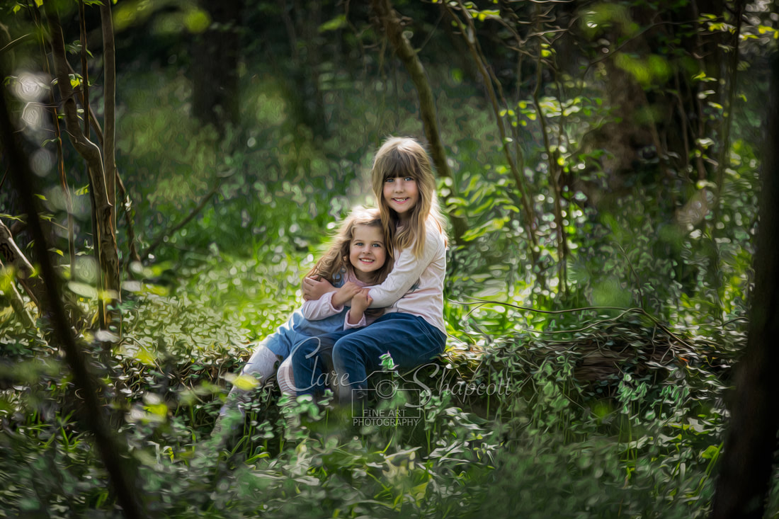 Two young girls sitting on log in the woods hold each other and smile.