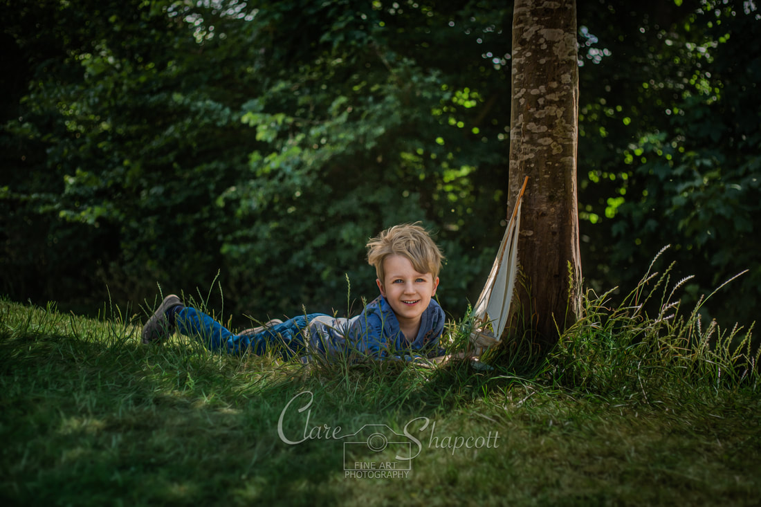 Young boy lying on the grass next to a tree smiles as he plays with wooden toy boat.