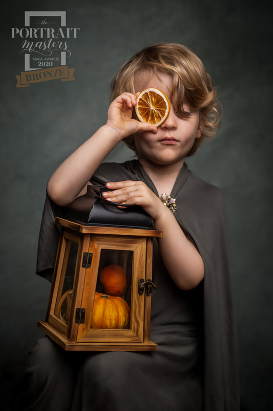 Young boy in grey cloak holds lantern filled with small pumpkins on lap whilst also holding a dried orange slice to his eye.