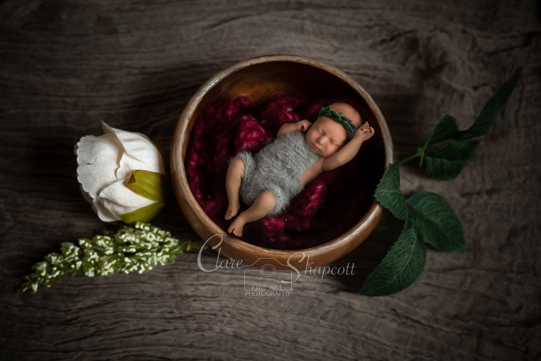Newborn baby sleeps on purple red fabric in large wooden bowl with leaves and flowers surrounding it.