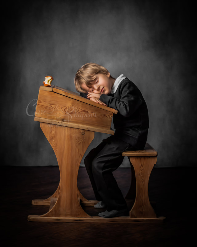 Young boy rests arms and head on traditional wooden school desk as he falls asleep with half eaten apple on the desk.