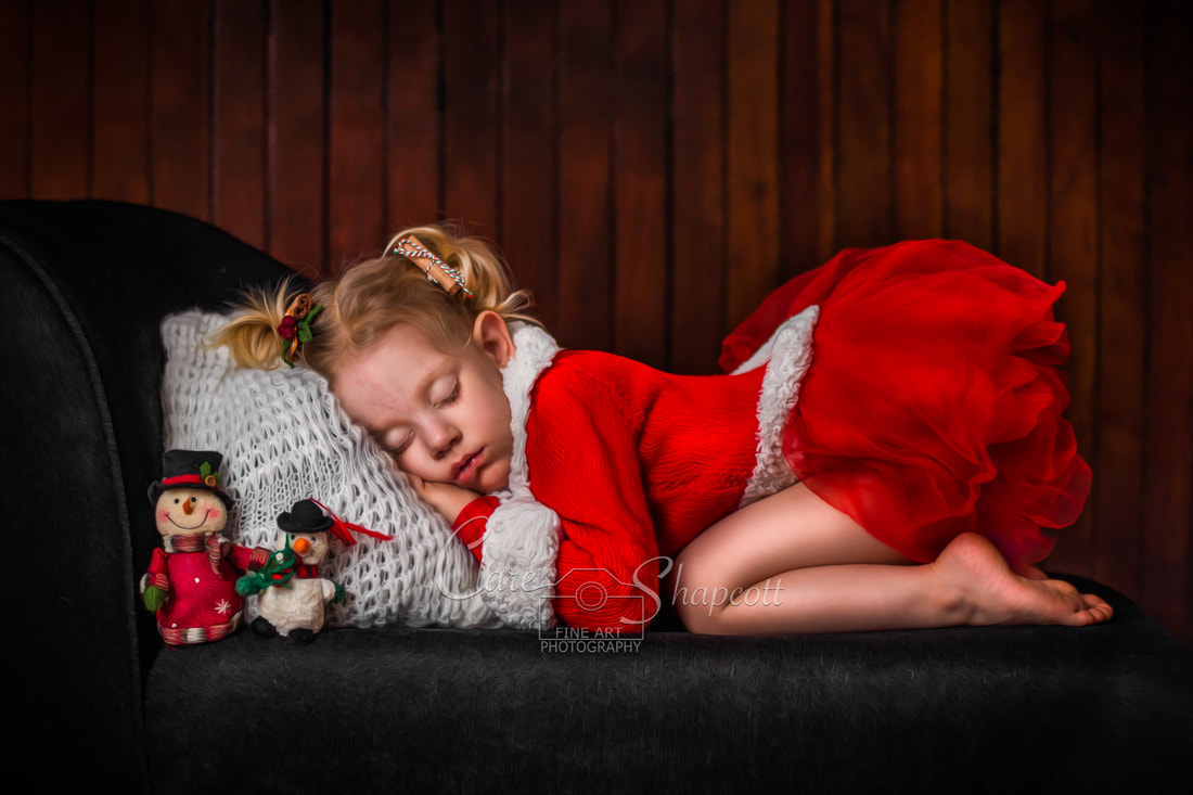 Young girl in red and white christmas cardigan sleeps on black chaise with two small snowman toys next to her.