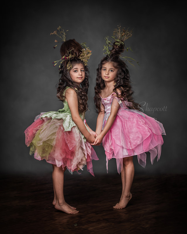 Two young girls hold hands as they are wearing pink and green tulle dresses, with small plants and flowers sticking out of their tall hairpieces. 