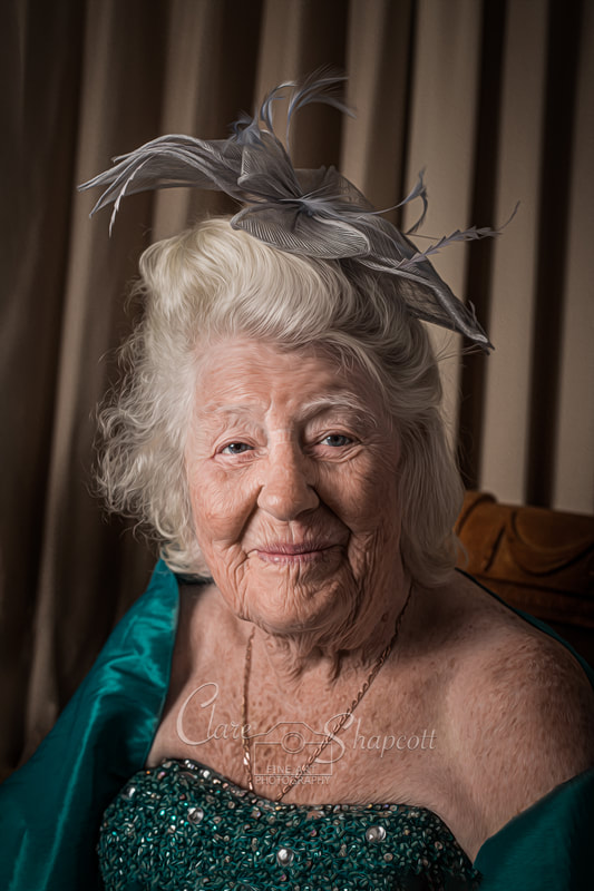 Elderly woman with beautiful grey headpiece and turquoise dress sits on brown chair and smiles at camera.