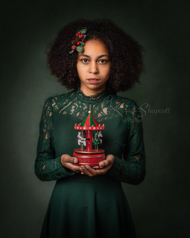 Curly haired girl in green dress with christmas bow in hair holds miniature christmas carousel in front of stomach.