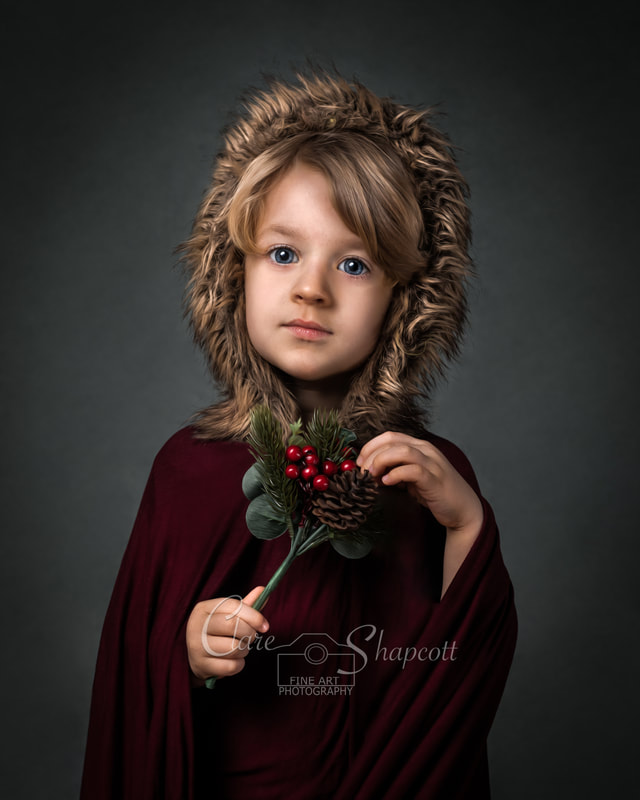 Young child in burgundy cloak and wool hood holds holly in front of grey background.