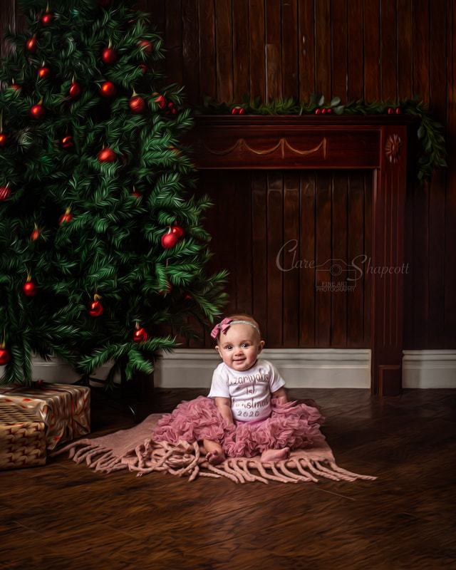 Portrait of baby sitting on pink material on the floor with a pink headband sitting next to a christmas tree with red baubles and presents.