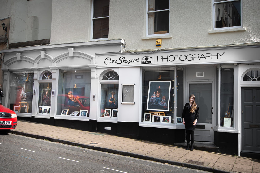 Clare stands in front of the door of her studio on the hill, various fine art photographs are displayed in the windows.