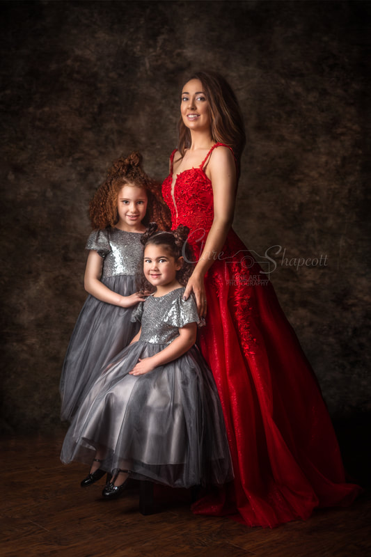 Mother in red sparkly dress stands next to her two curly haired daughters wearing silver sparkly dresses in front of a brown background.