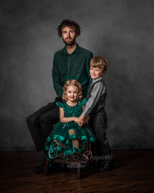 Man wearing smart green shirt sits on stool in front of daughter in green dress, and son in grey suit.