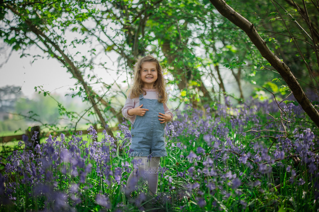 Young girl in blue dungarees stands among bluebell flowers as she smiles at camera.