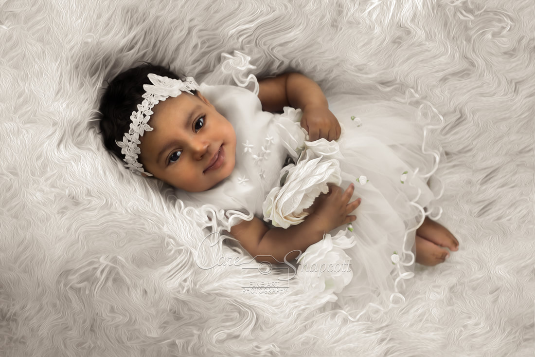 Baby in white dress and white flower headband lays on soft white material and smiles upwards at camera.