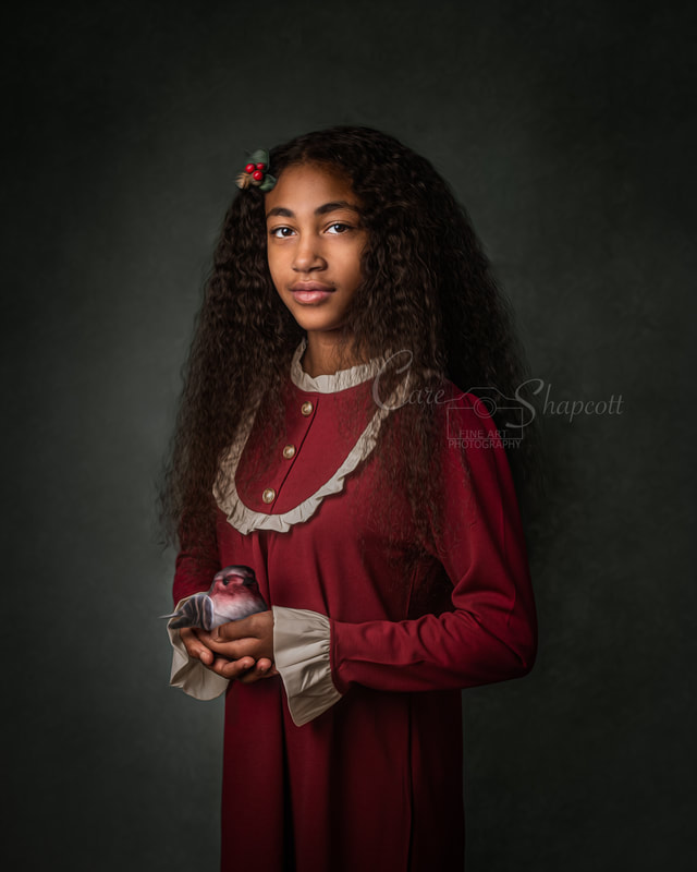 Girl with long curly hair in long red gown stands with holly hair clip in her hair whilst holding a robin ornament.