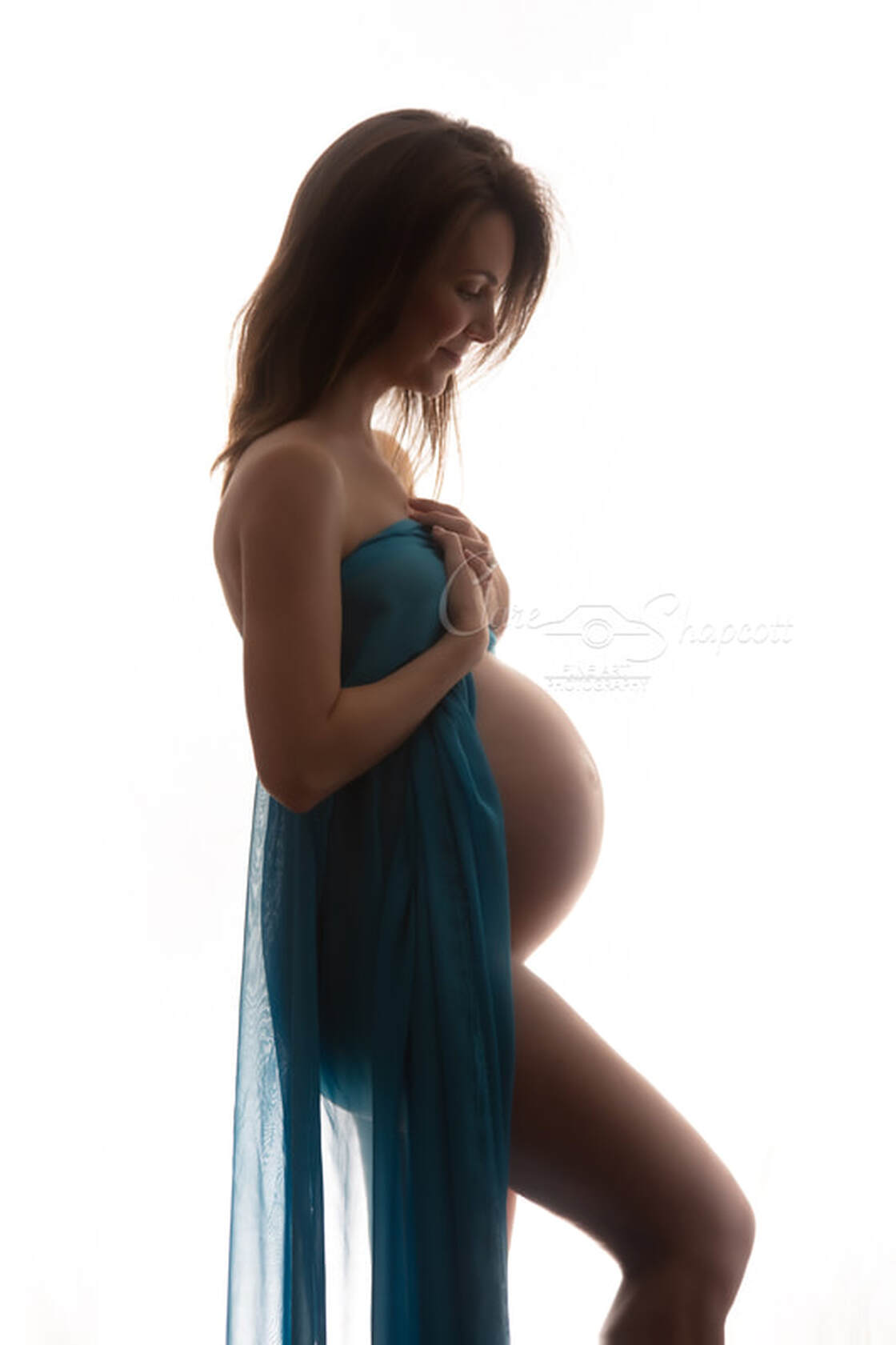 Woman with brown hair holds light blue material over herself as it droops to the floor during nude maternity photoshoot.