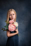 Fine art portrait of young blonde girl in blue dress holding pink and white flowers in front of blue background.