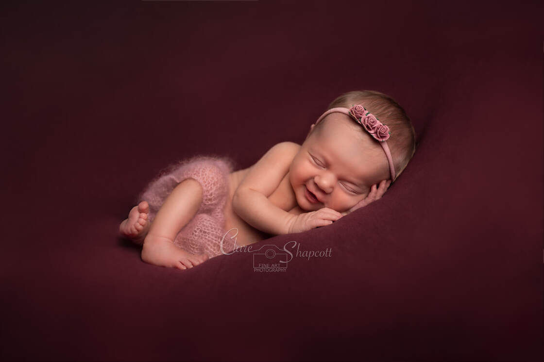 Sleeping newborn with soft pink trousers lies down on purple material while wearing a pink flower headband